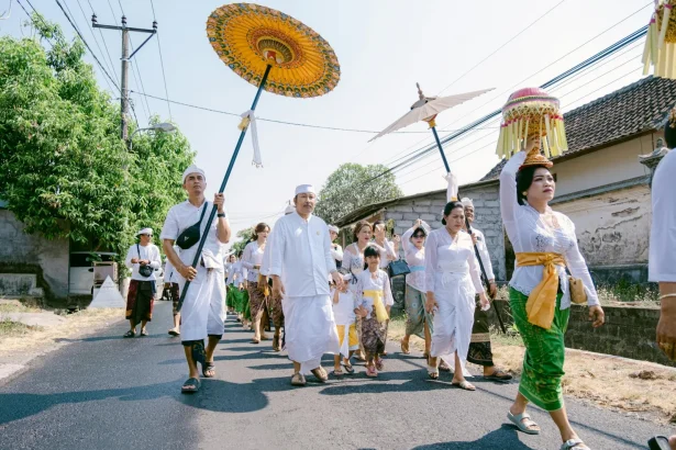 A Balinese procession with traditional attire and vibrant umbrellas on a sunny day.