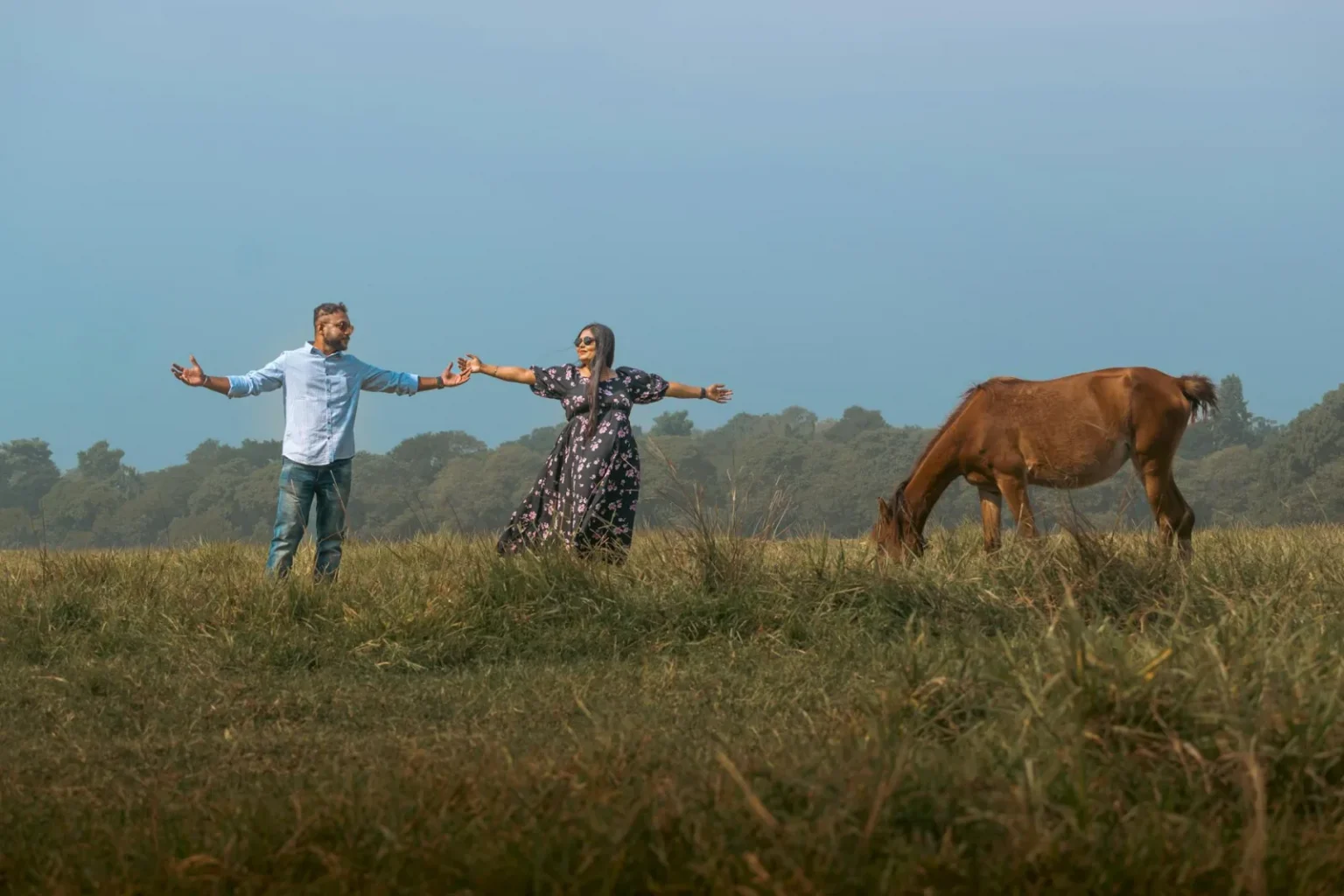 Couple holding hands in scenic Kolkata field with a horse grazing nearby.