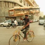 A man rides a bicycle balancing a tray of bread on his head in a busy city street.