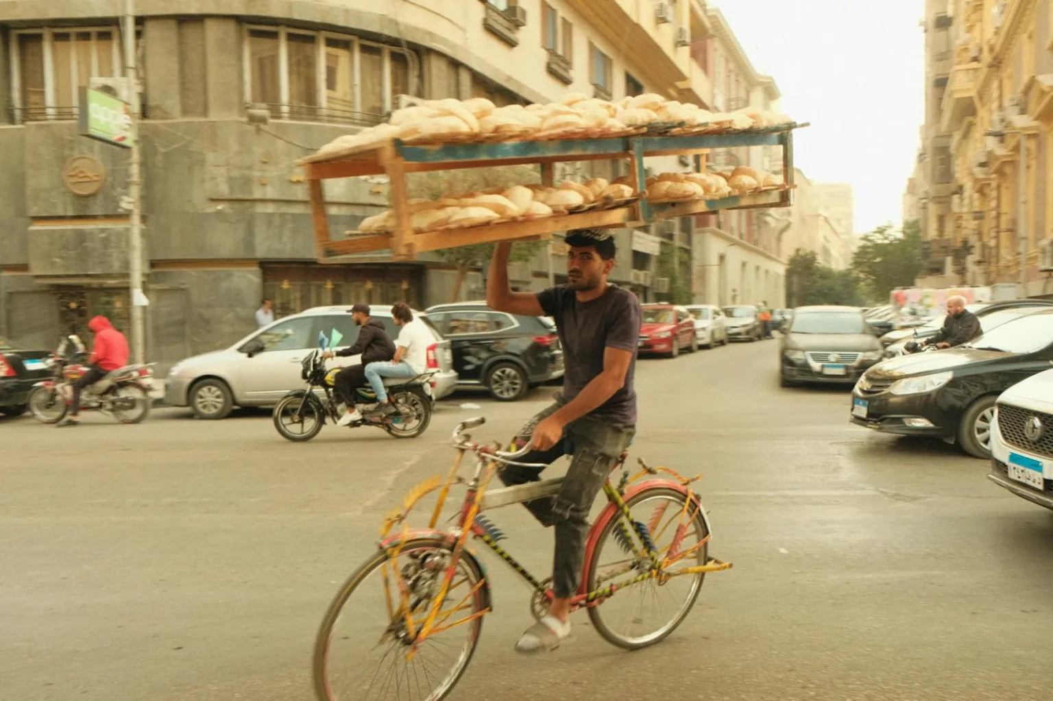 A man rides a bicycle balancing a tray of bread on his head in a busy city street.