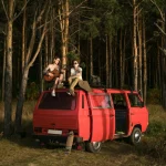 Two adults enjoy a serene moment atop a red van beside a lush forest.