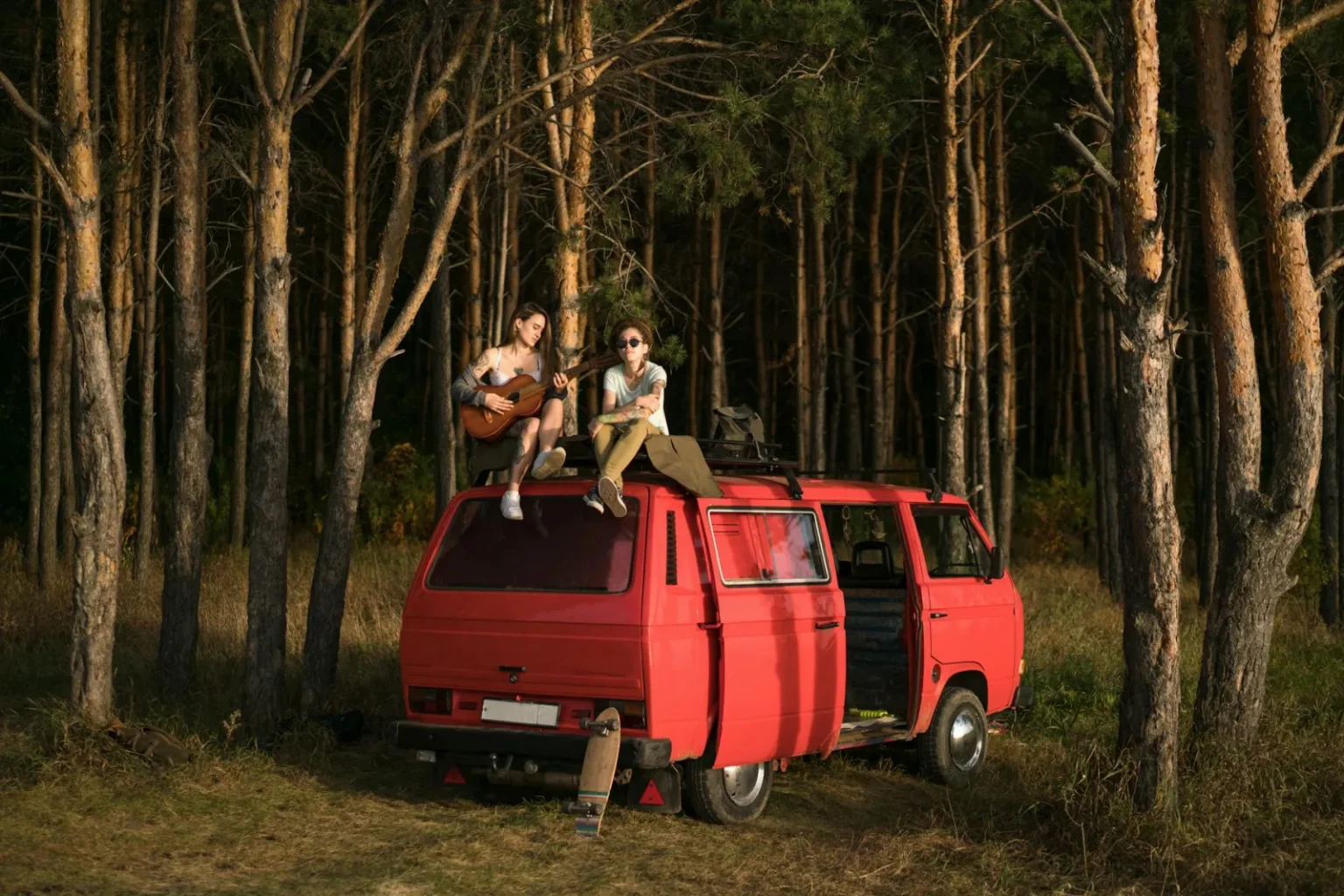 Two adults enjoy a serene moment atop a red van beside a lush forest.