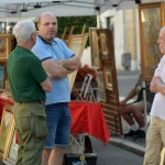 Three men conversing at an outdoor art market in Villafranca di Verona, Italy.