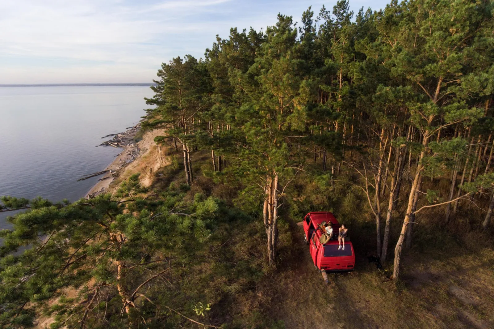 Drone captures a red van parked near a forest cliff overlooking a serene coastline.