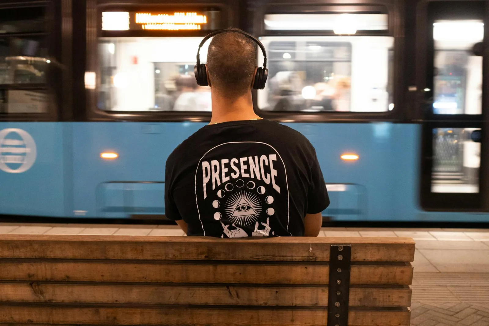 A man with headphones sits on a bench as a bus passes by in a city setting.