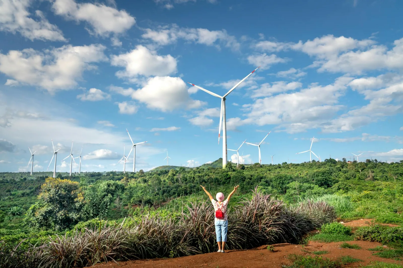 A woman joyfully raises her arms in front of wind turbines on a lush green hillside under a blue sky.