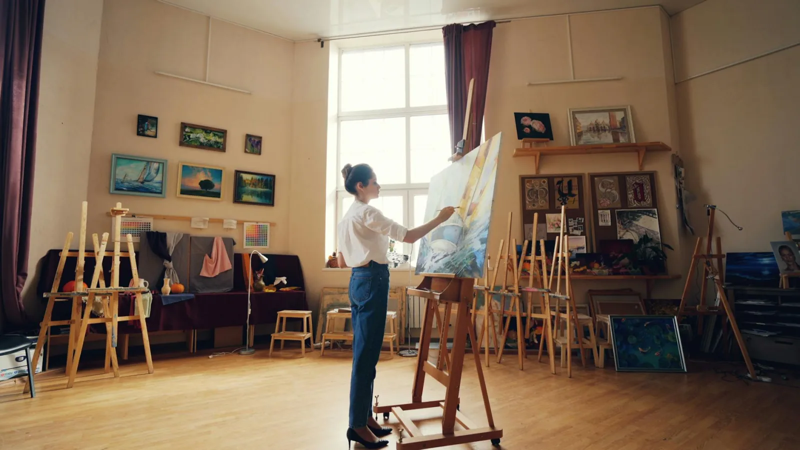 A female artist in a sunlit studio paints on a canvas. Artworks decorate the walls.