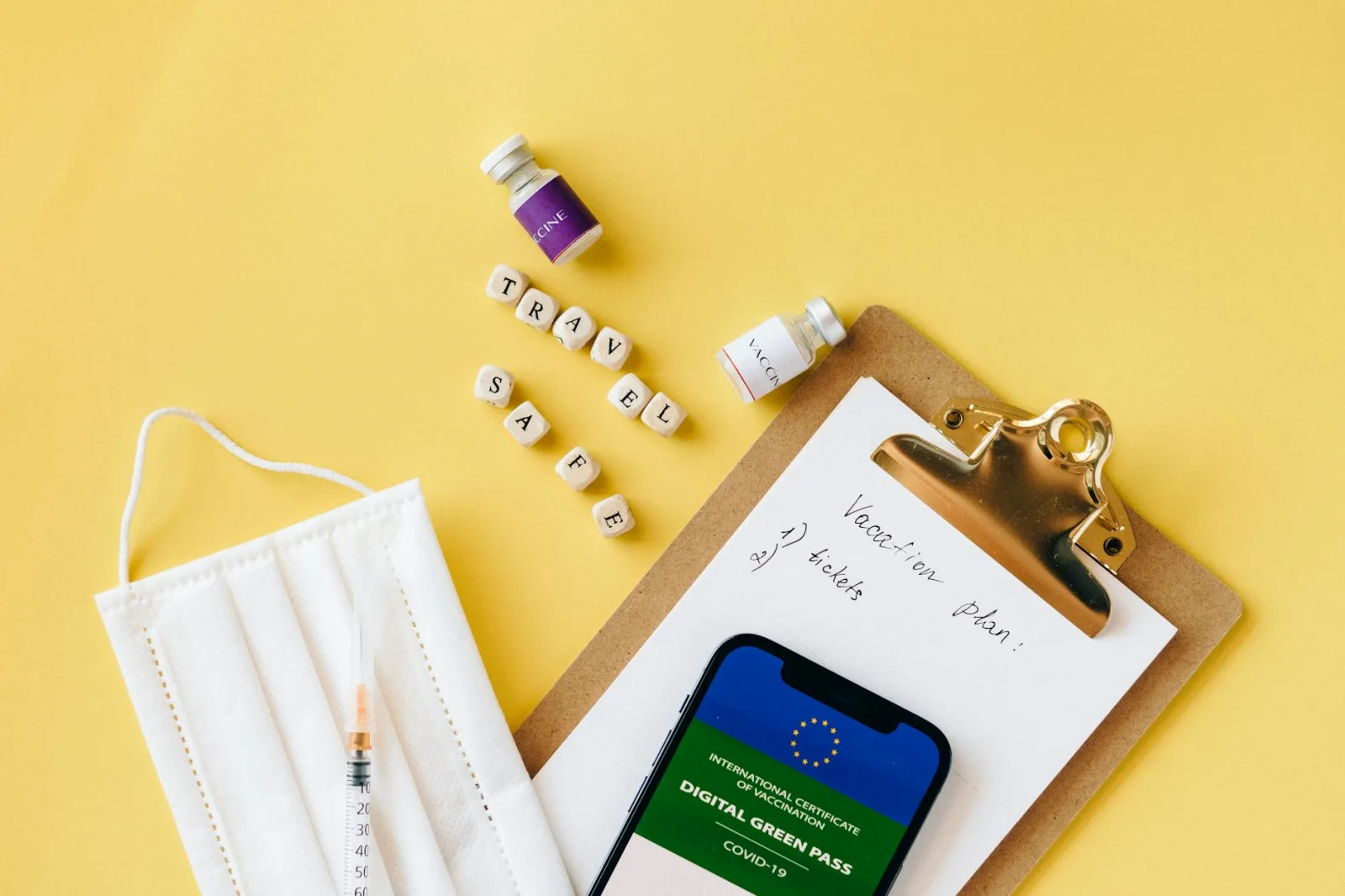 Flat lay of travel safety items: vaccine, mask, digital green pass on a vibrant yellow background.