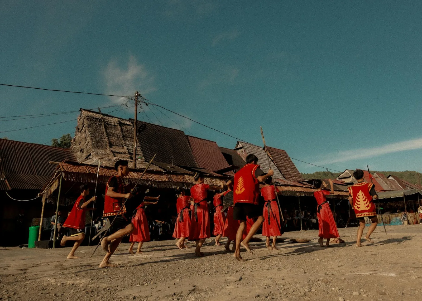 Men performing a traditional dance in a rural village, dressed in vibrant red attire under a clear sky.