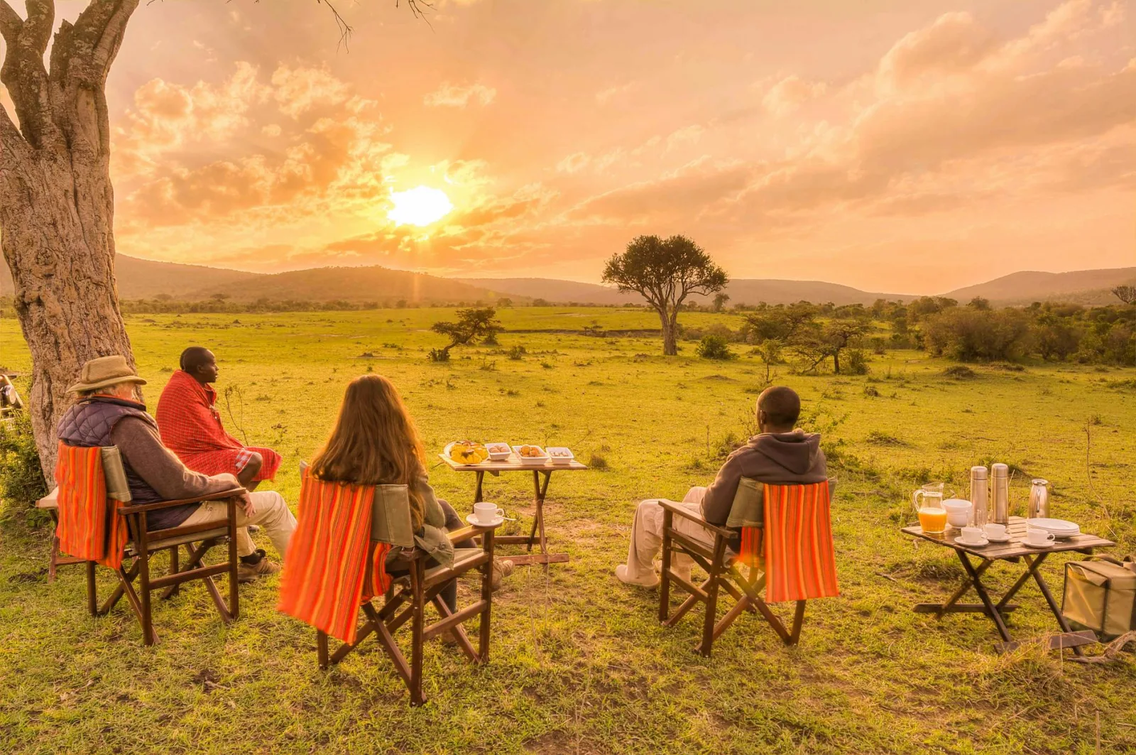 Four people enjoy a sunset picnic in the African savanna, surrounded by nature and serene views.