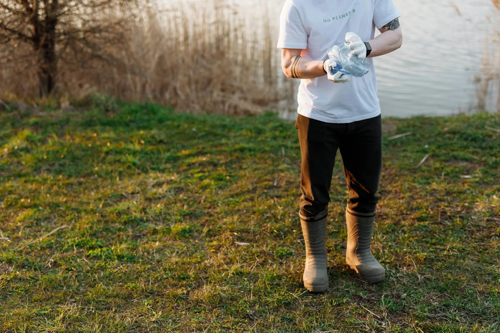 An adult in rubber boots picks up trash by a lake, promoting environmental conservation.