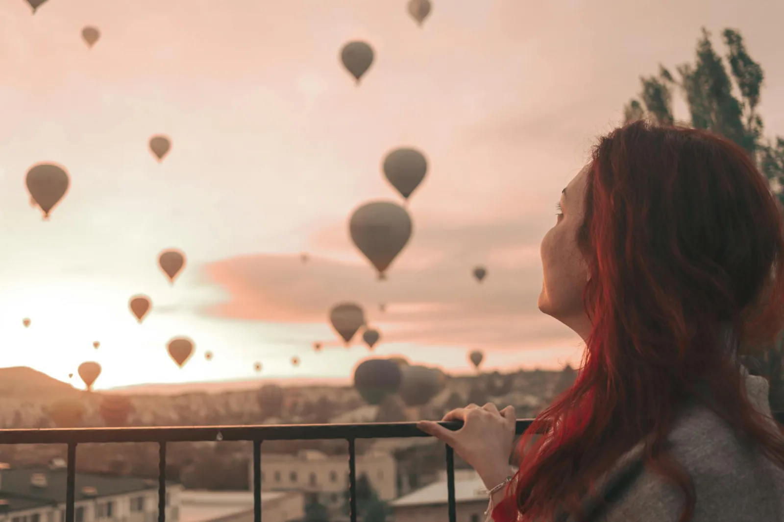 A woman with red hair watches hot air balloons soar over Cappadocia during a serene sunrise.