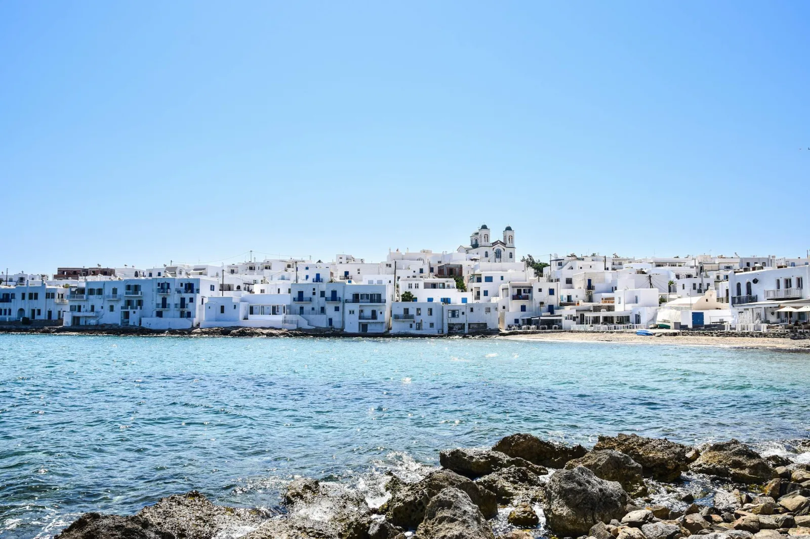 Whitewashed buildings and a church by the sea in a Greek town under a clear blue sky.