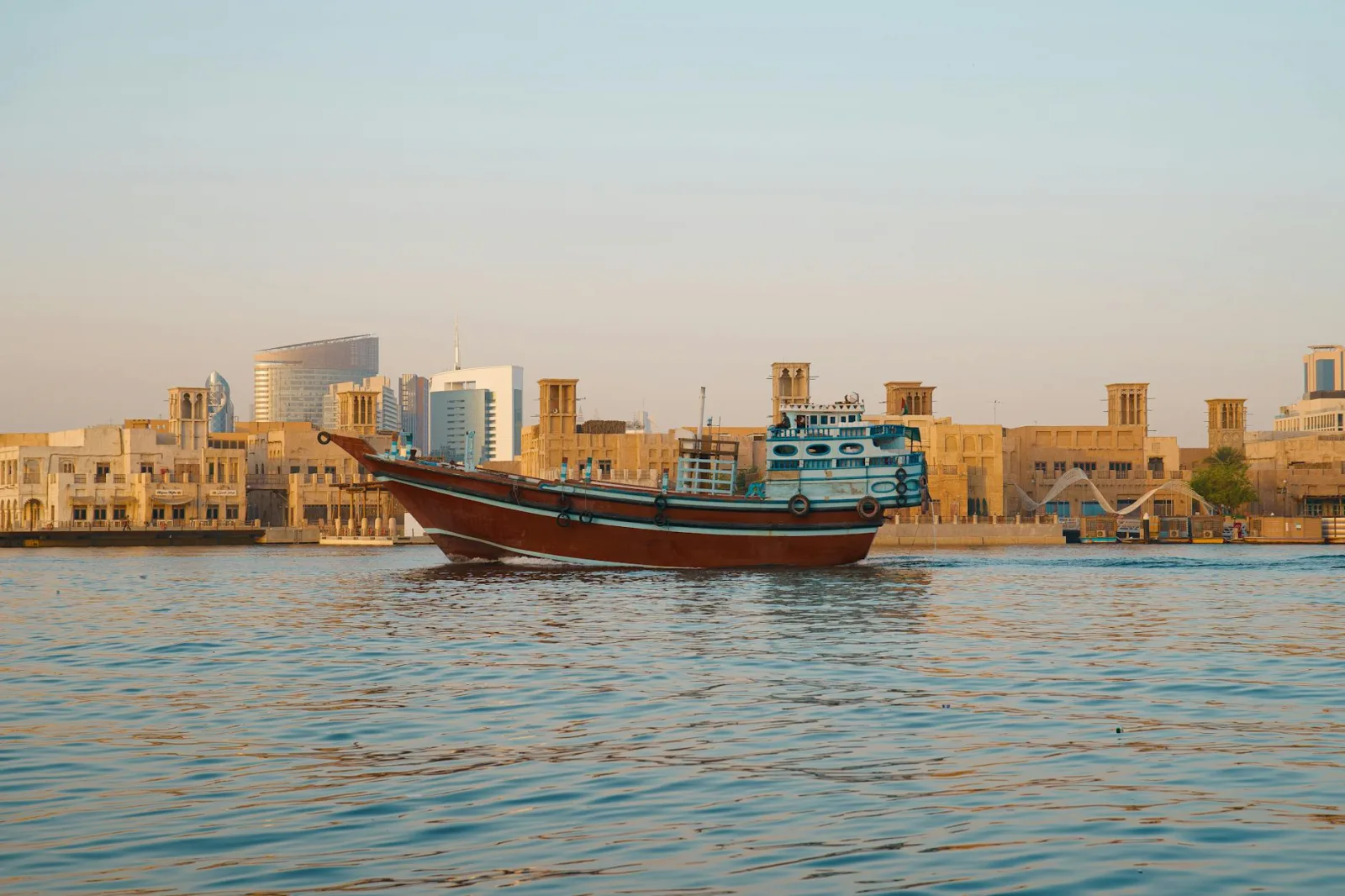 A traditional dhow boat sails through Dubai Creek with iconic architecture in the background.