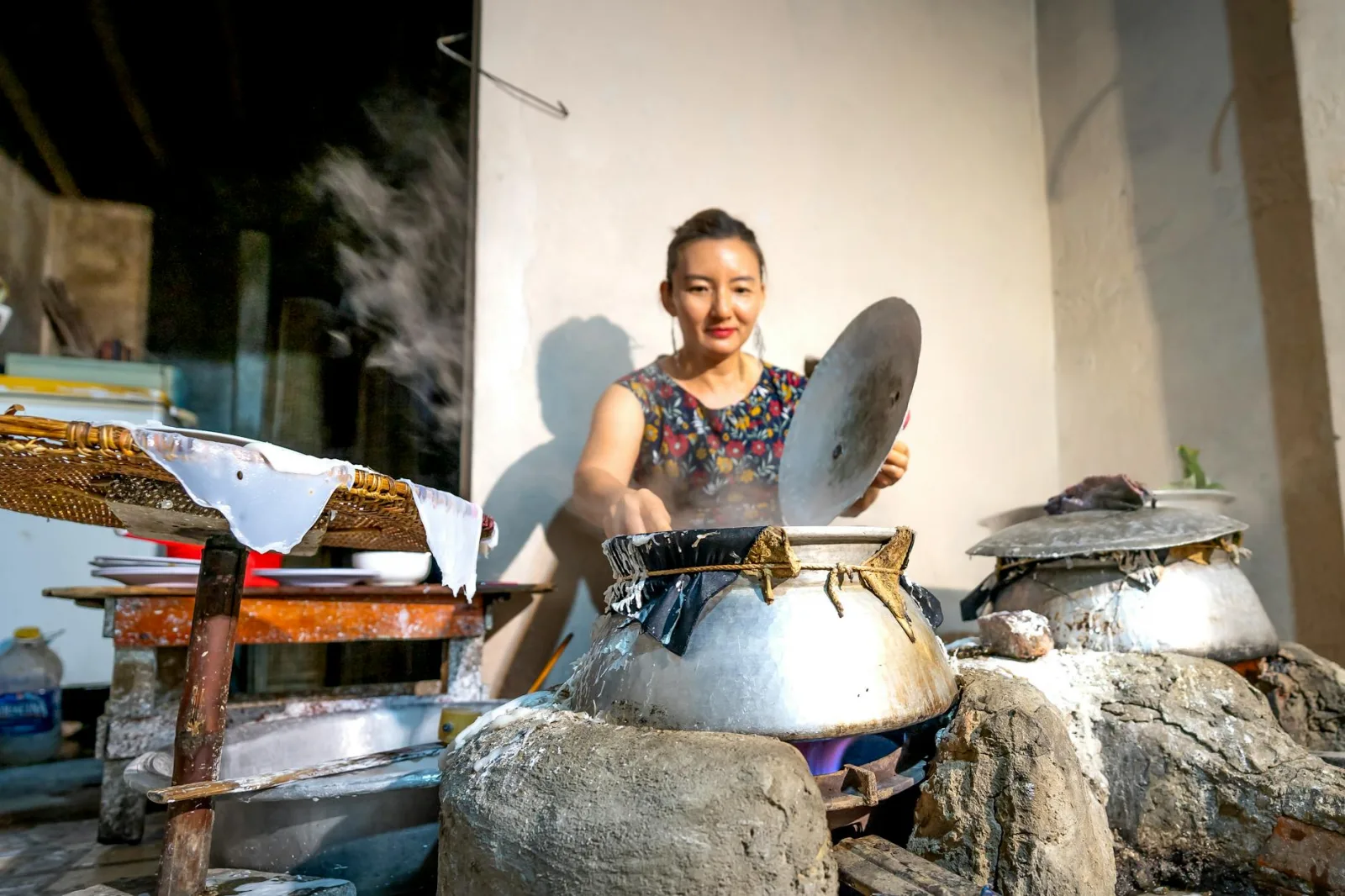 Asian woman cooking in traditional outdoor kitchen with steaming pots. Rustic and cultural scene.