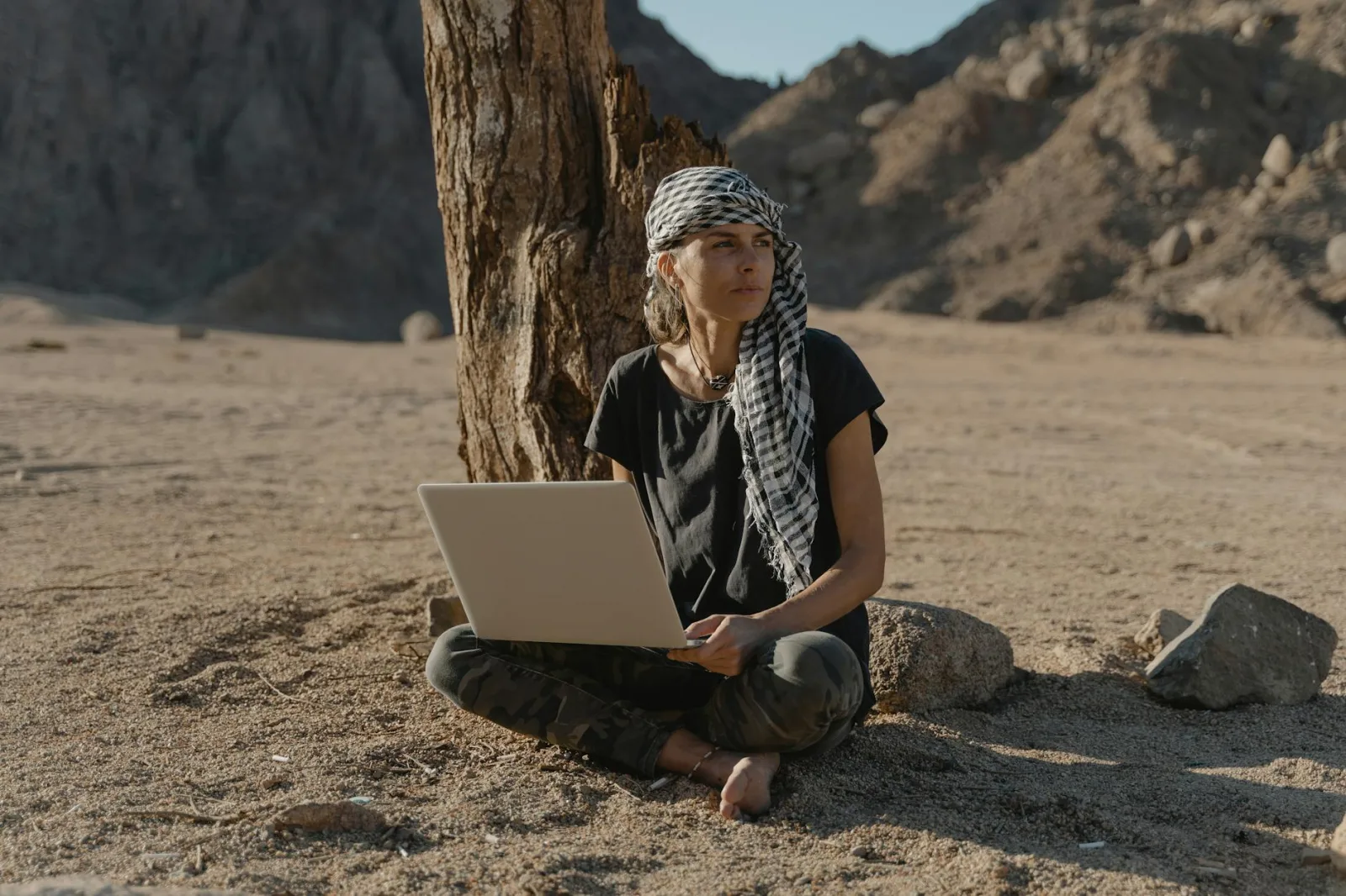 Caucasian woman sitting barefoot with laptop under tree in arid landscape.