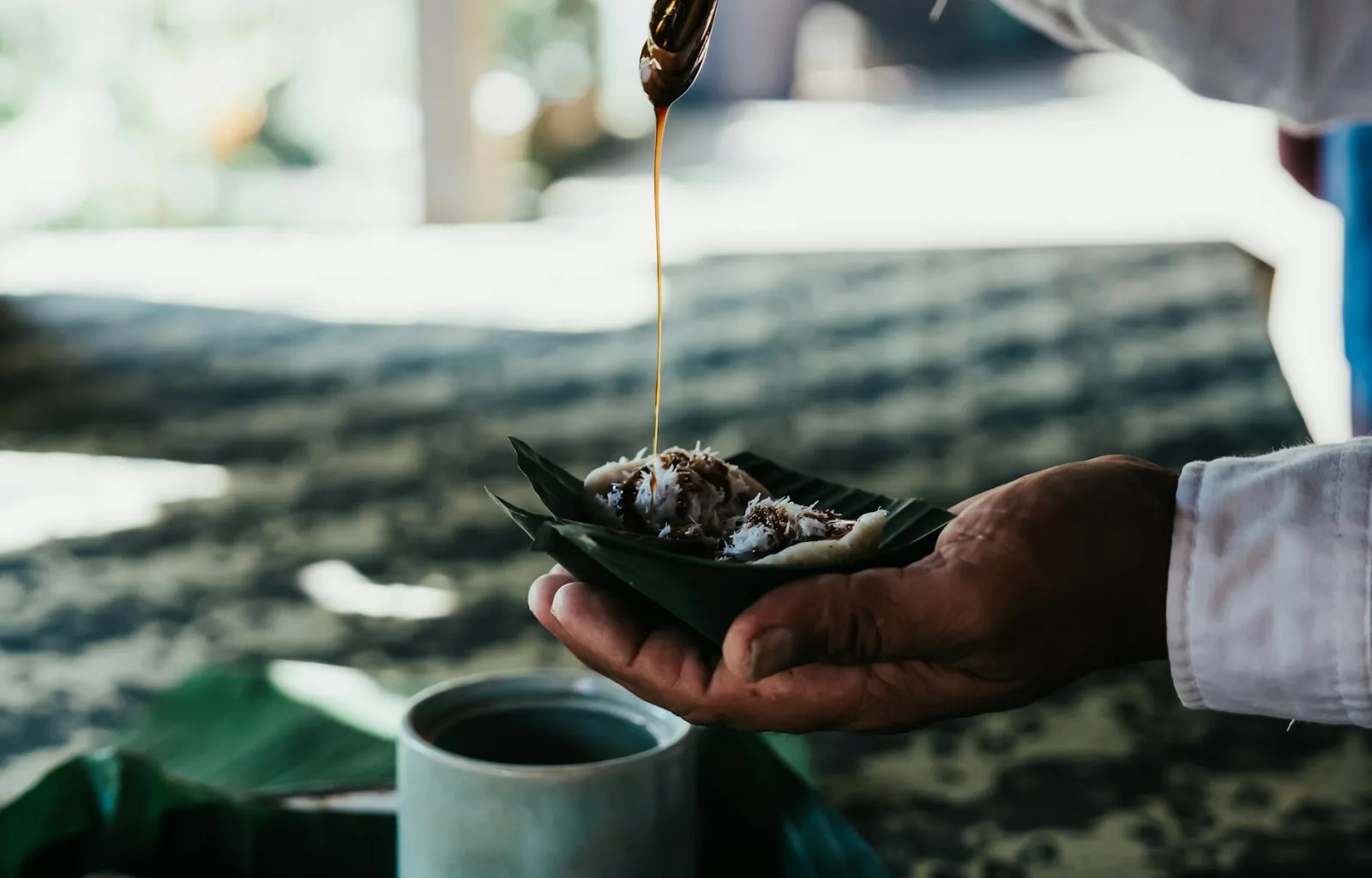 A hand holding traditional food with sauce drizzle over banana leaf captured outdoors.