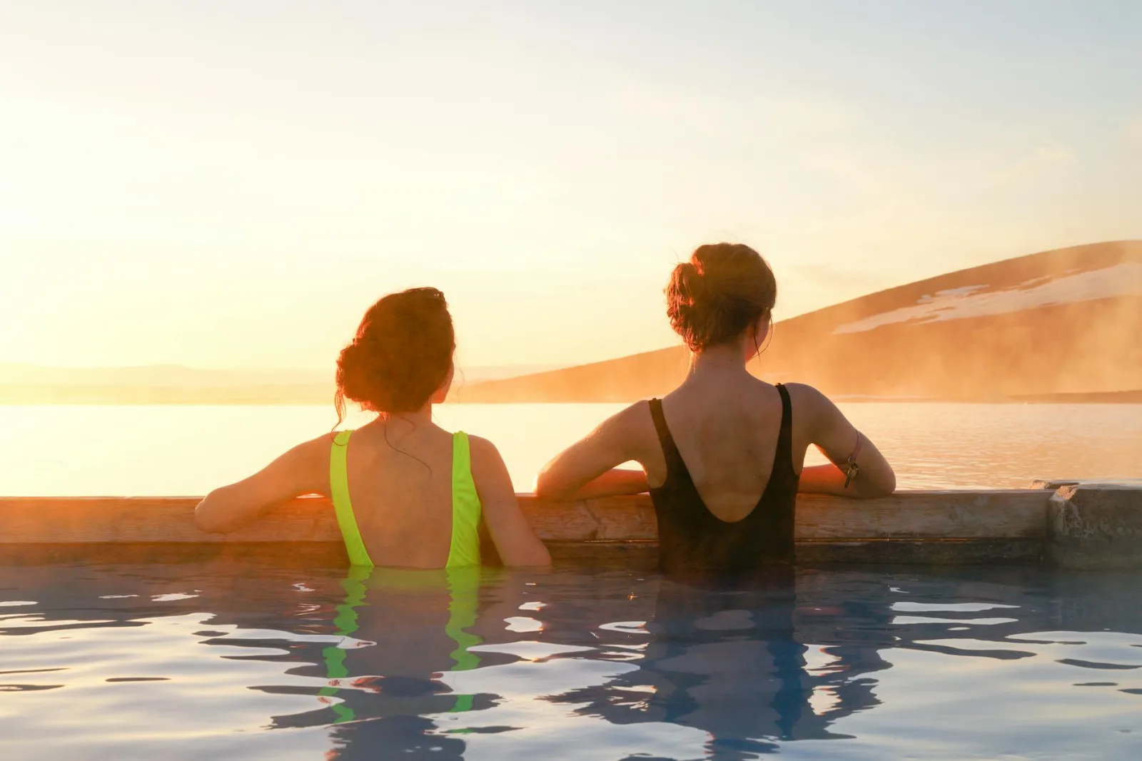 Two women relaxing in a hot spring, enjoying the serene sunrise view over the mountains.
