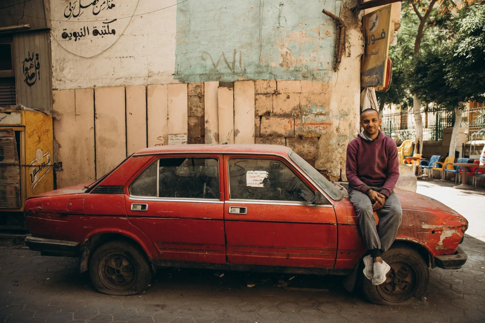 A man casually sitting on a red vintage car in a Cairo street, exuding an urban vibe.