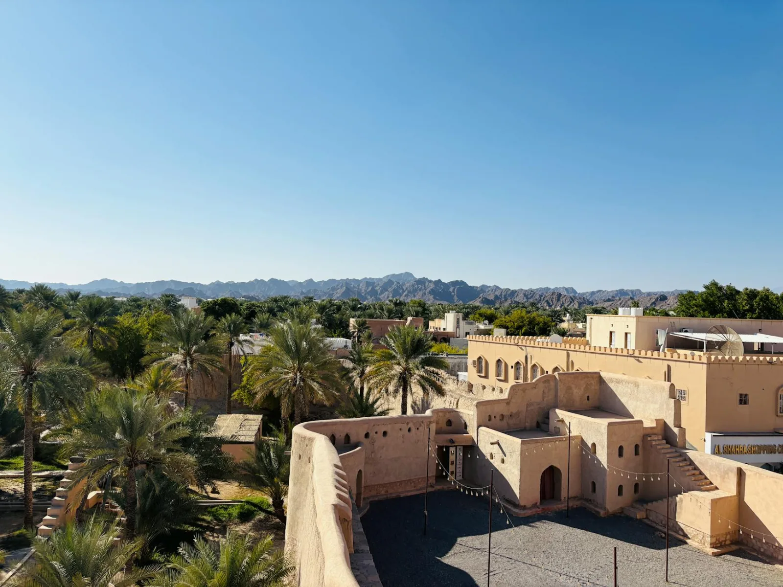 View of Nizwa Fort and the surrounding oasis against Hajar Mountains under a clear blue sky.