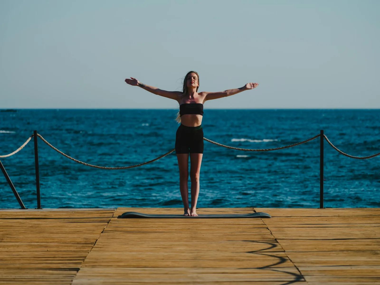 A woman in activewear stands in a yoga pose on a wooden dock near the sea, embracing a tranquil moment.