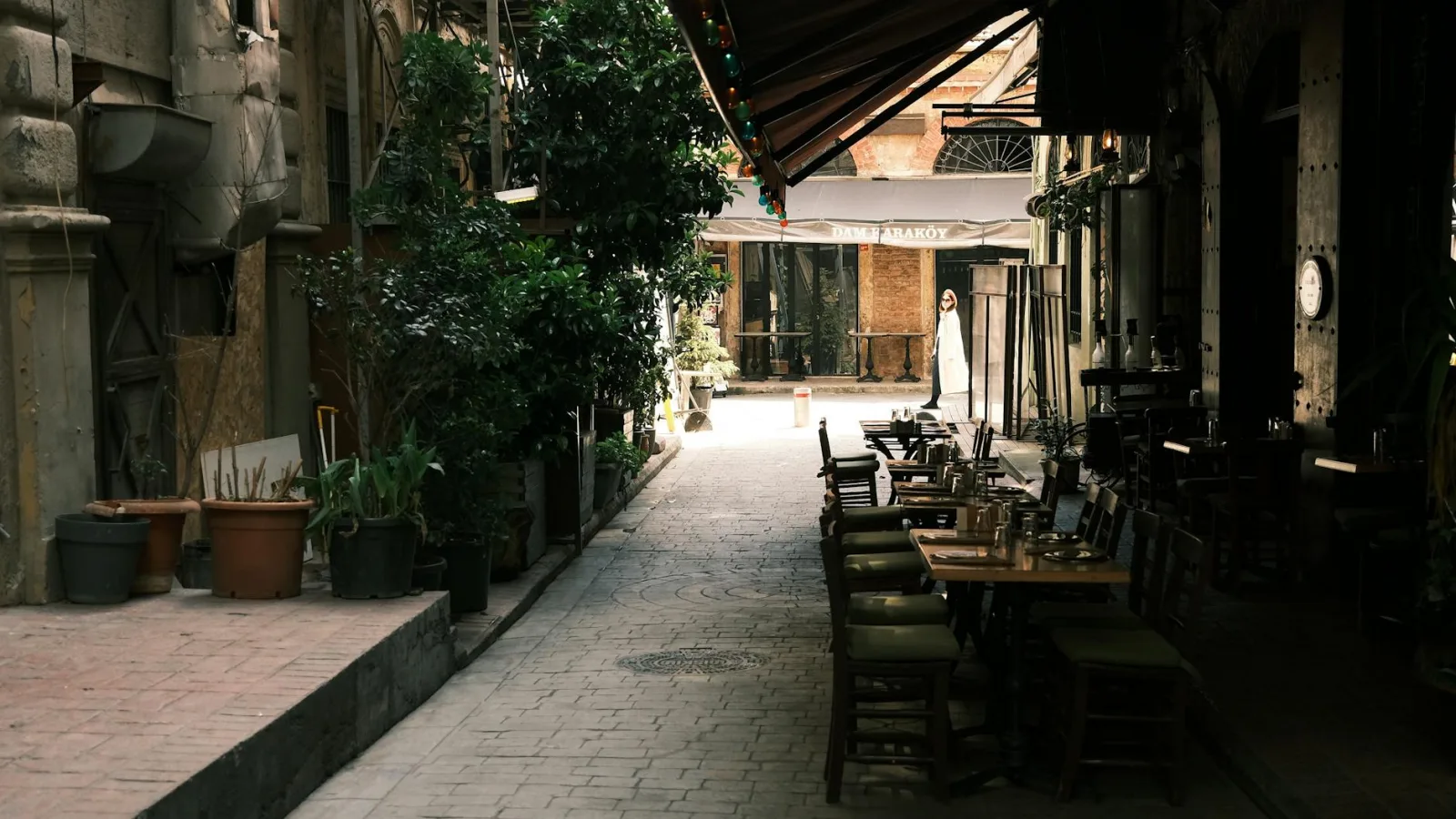 Cozy alleyway with outdoor cafe seating, lush greenery, and a solitary pedestrian.