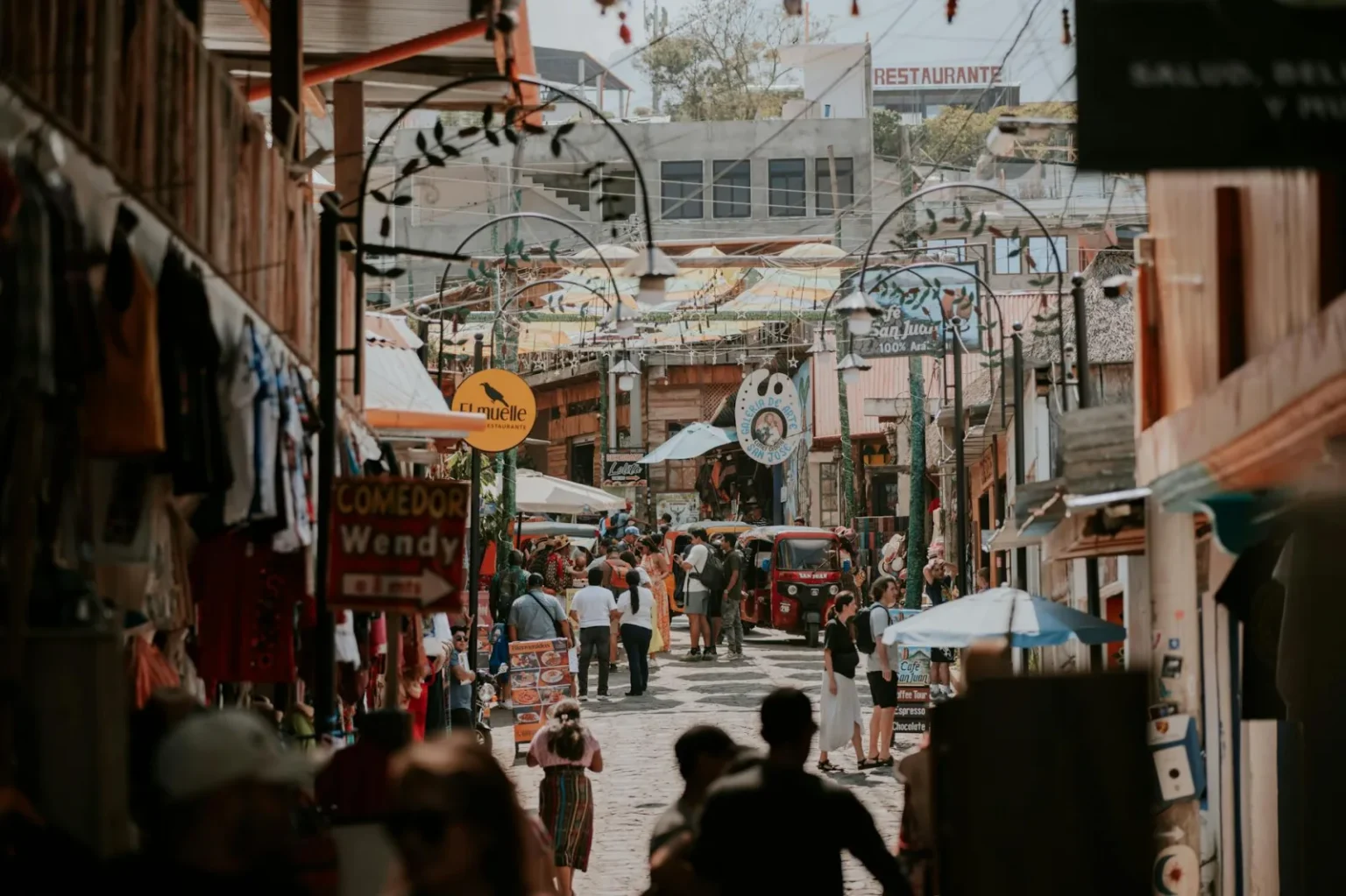 A lively market street filled with people and colorful shops in a sunny village.