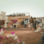 A festival scene at a beach resort with people, colorful flowers and a sign reading 'Obrigado'.