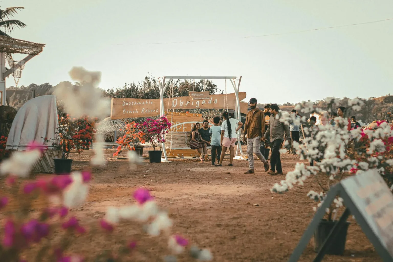 A festival scene at a beach resort with people, colorful flowers and a sign reading 'Obrigado'.