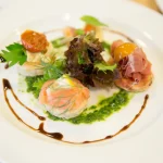 A beautifully plated gourmet dish featuring smoked salmon, prosciutto, and greens on a white plate.