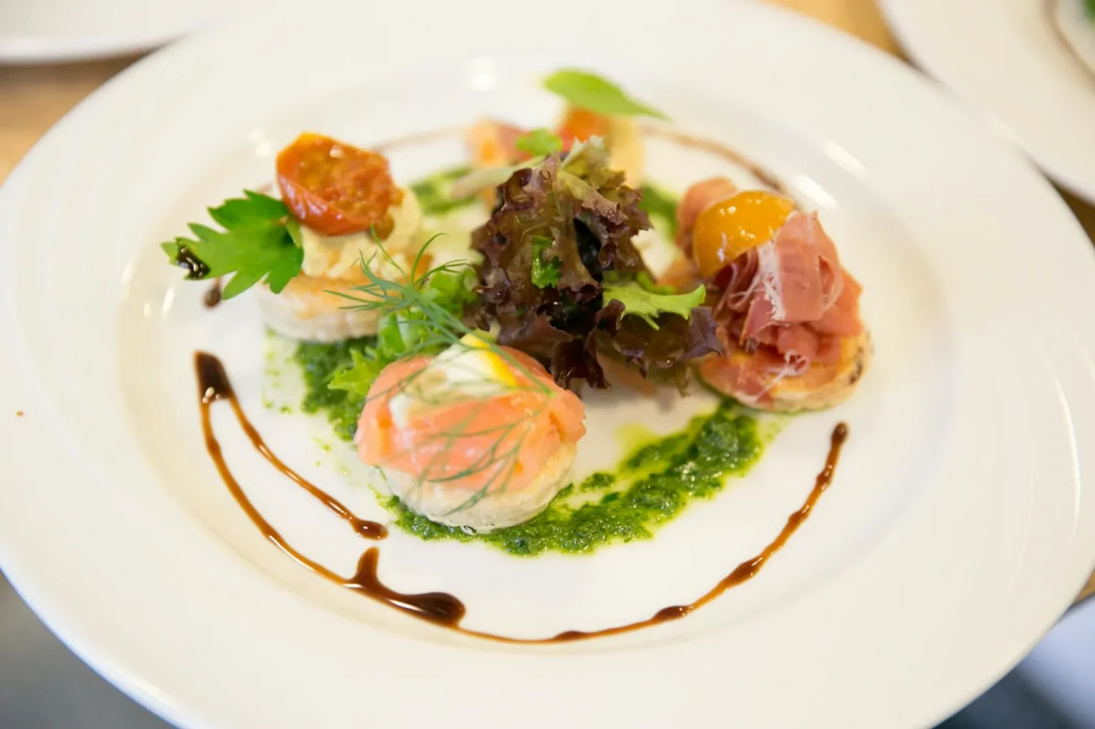 A beautifully plated gourmet dish featuring smoked salmon, prosciutto, and greens on a white plate.