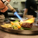 Searing gourmet dish with fresh vegetables using a culinary torch in a professional kitchen.