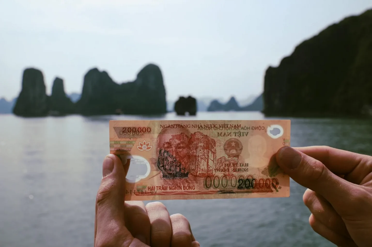 Hands holding Vietnamese Dong with Halong Bay in the background, a fusion of finance and travel.