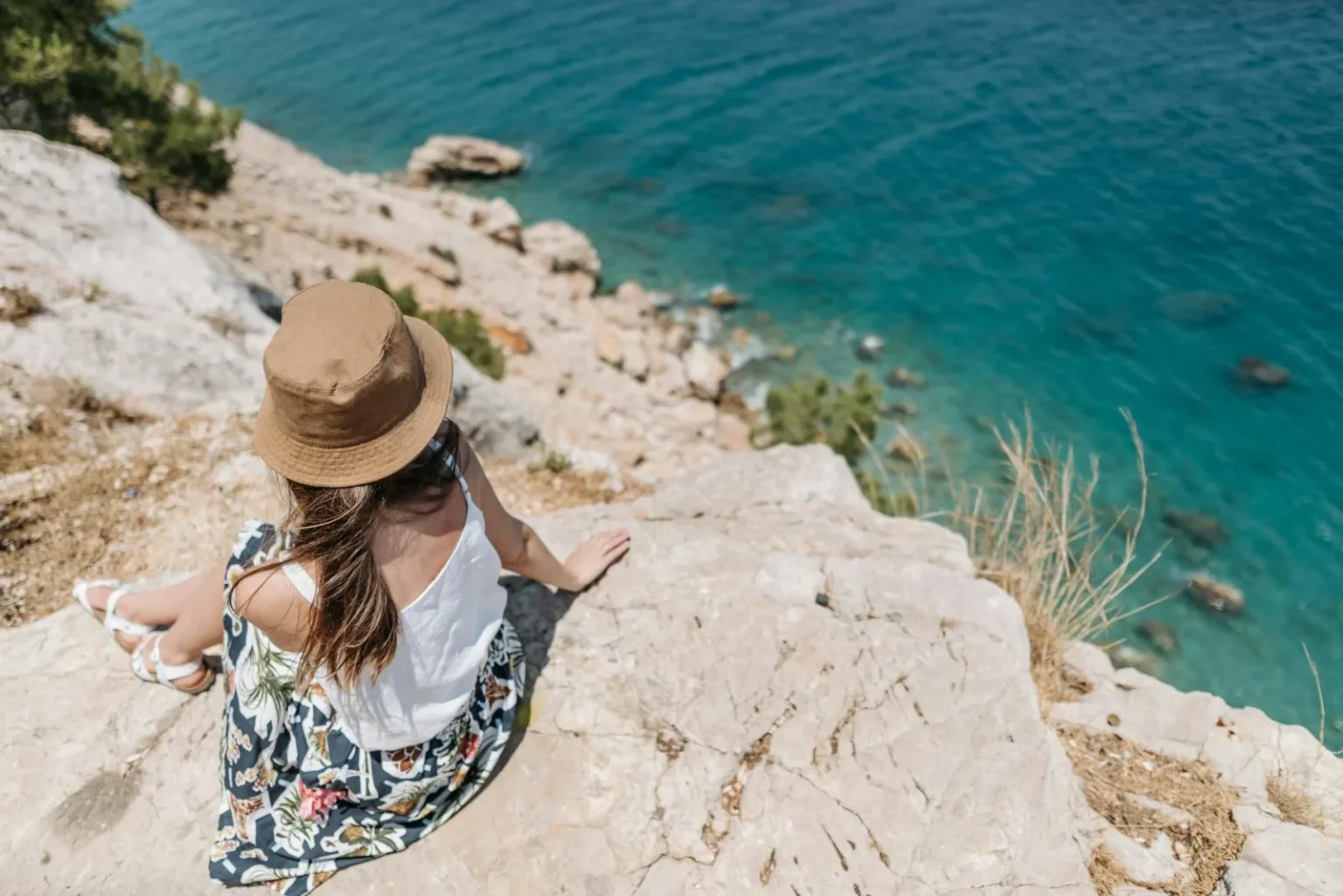 A woman sits on a rocky cliff, enjoying the summer view of turquoise waters.