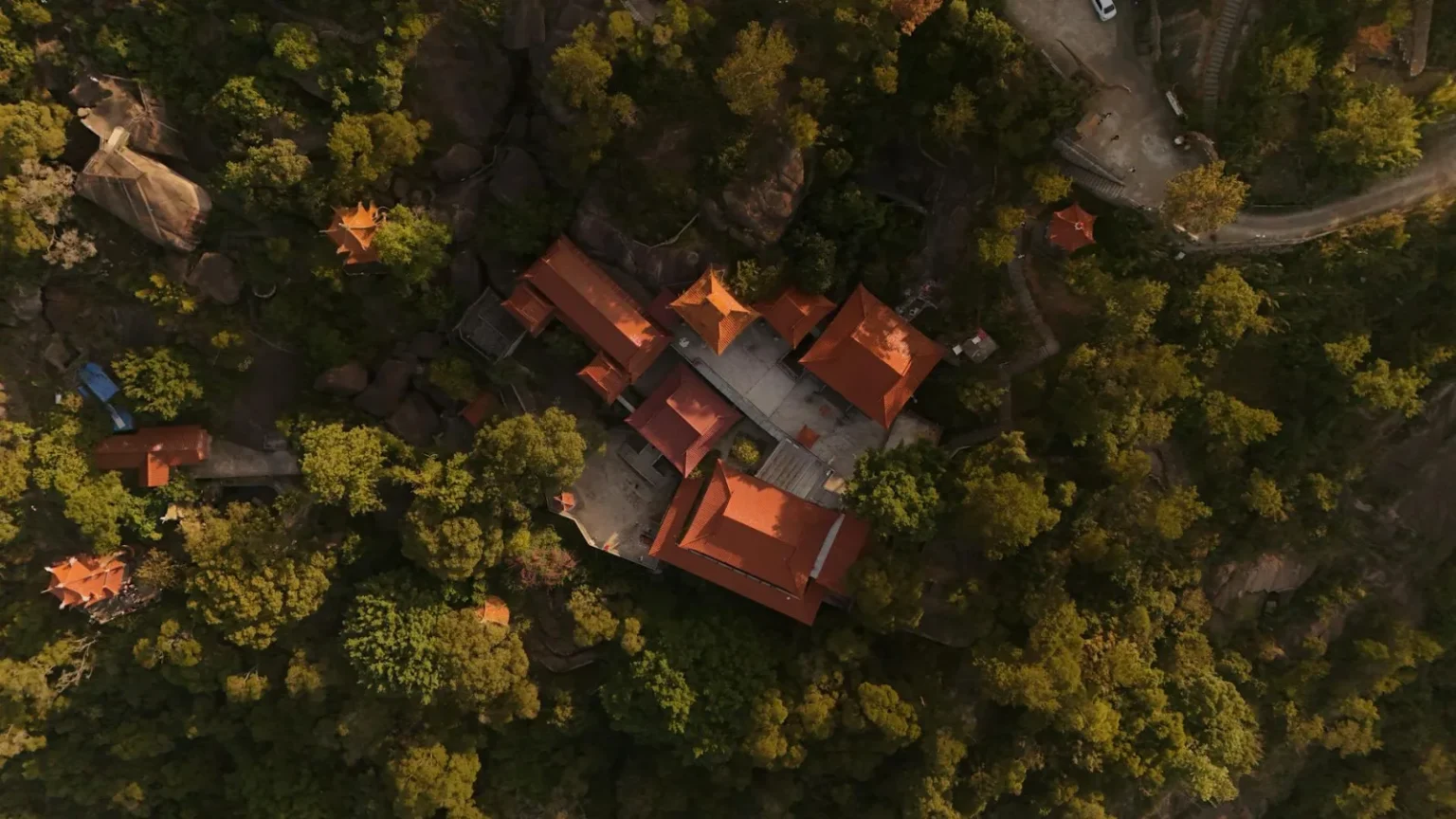 Aerial shot of a temple with red roofs surrounded by dense forest on a hilltop.