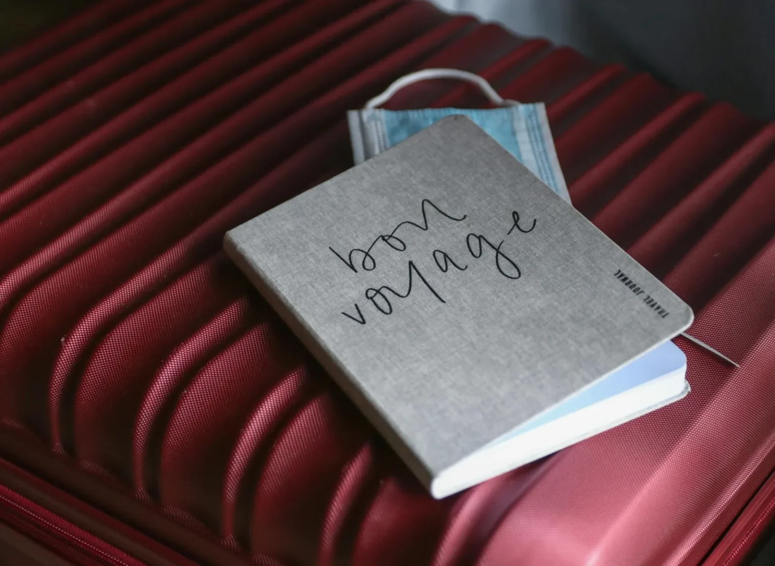 Close-up of a travel journal and mask on a red suitcase, symbolizing preparation for a voyage.