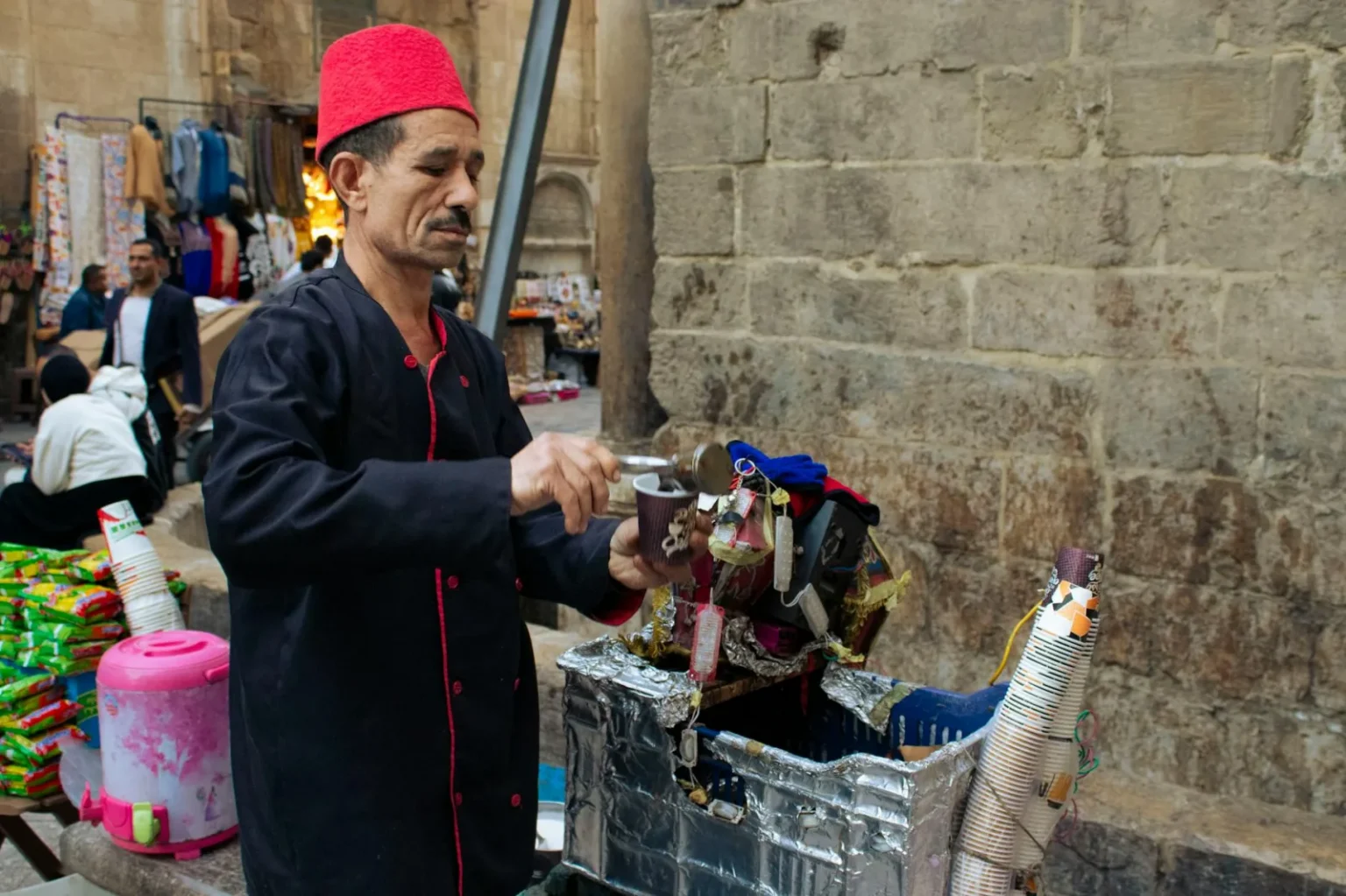 A street vendor in a red fez prepares a traditional drink at a bustling market.