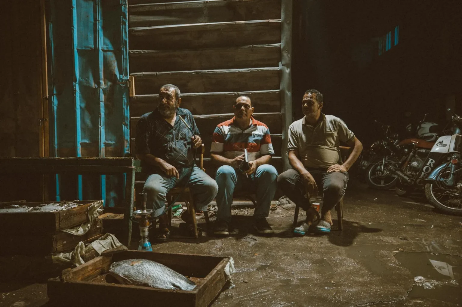 Three men sitting at a fish market in Ras El-Bar, Egypt, with motorcycles in the background.