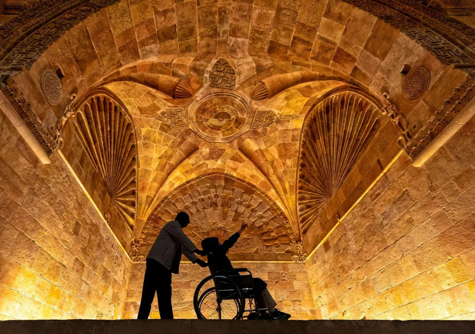 Silhouette of a man assisting a person in a wheelchair under a historic, ornately decorated arched ceiling.