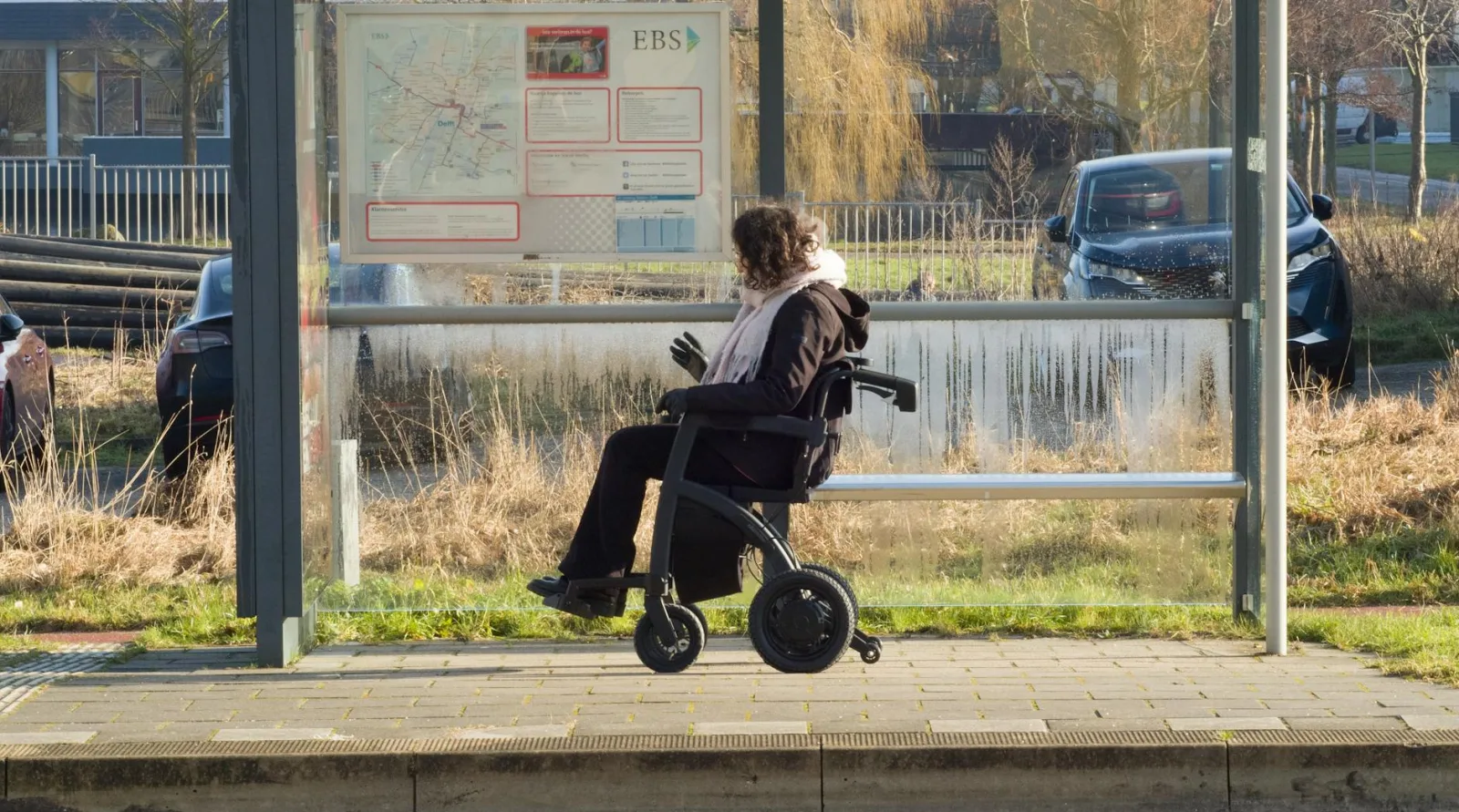 A woman in a wheelchair patiently waits at a bus stop in Delft, Netherlands on a chilly day.