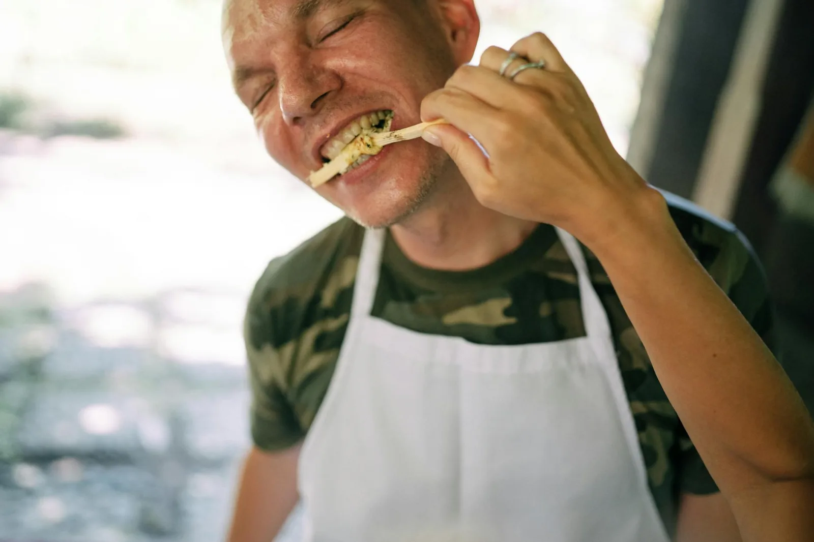 Man wearing a camo shirt and apron savoring food outdoors with an intense expression.