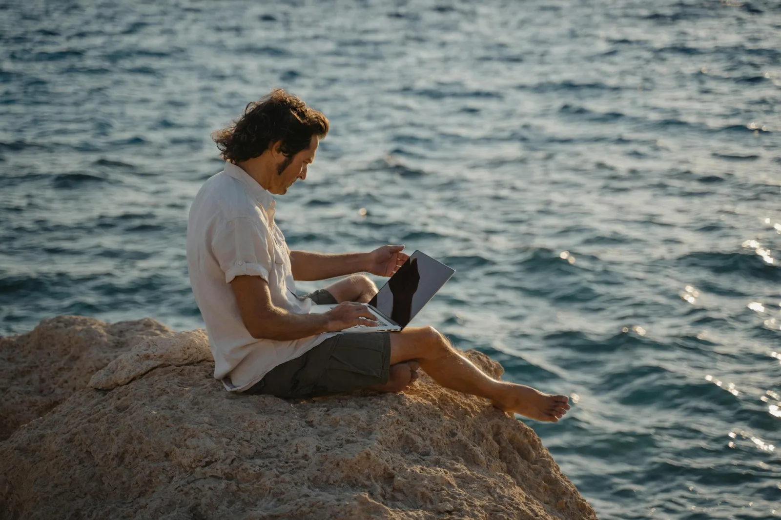A man sitting on rocky terrain by the sea, working remotely on his laptop during the day.