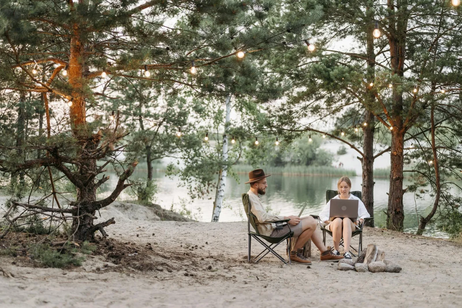 A couple enjoys a serene camping experience by a lake, working on a laptop under string lights.
