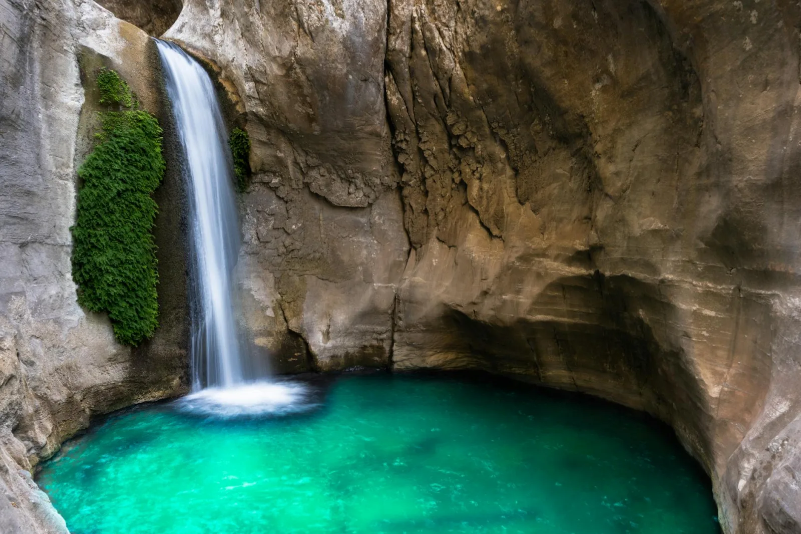 A beautiful waterfall flows into an emerald pool inside a rocky cave, creating a serene natural scene.