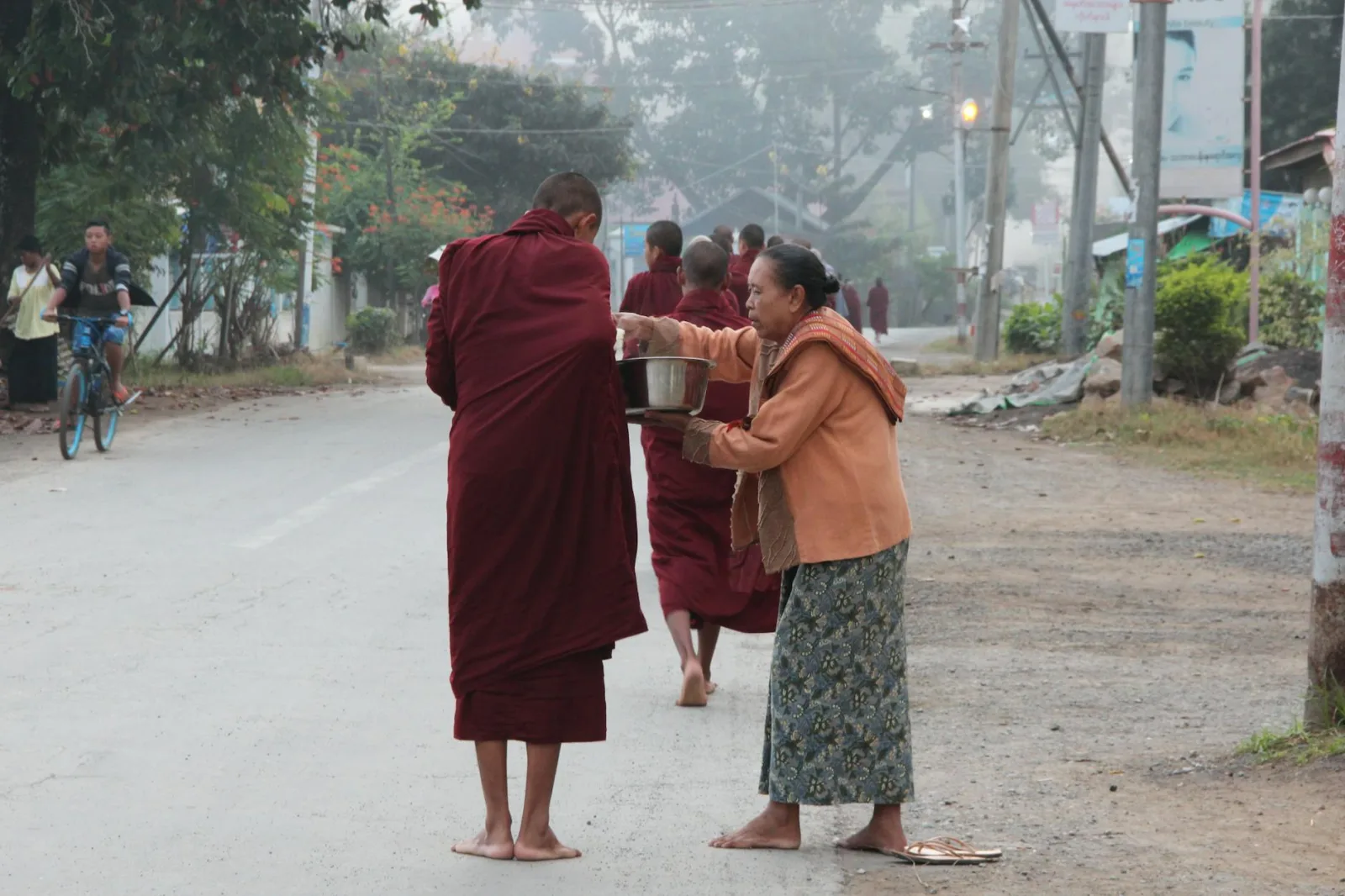 A woman offers alms to a Buddhist monk on a street in Shan, Myanmar, showcasing cultural traditions.