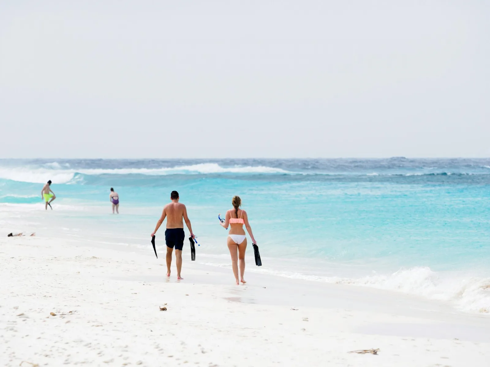A man and woman walk along a serene beach, carrying snorkeling gear by the turquoise ocean.