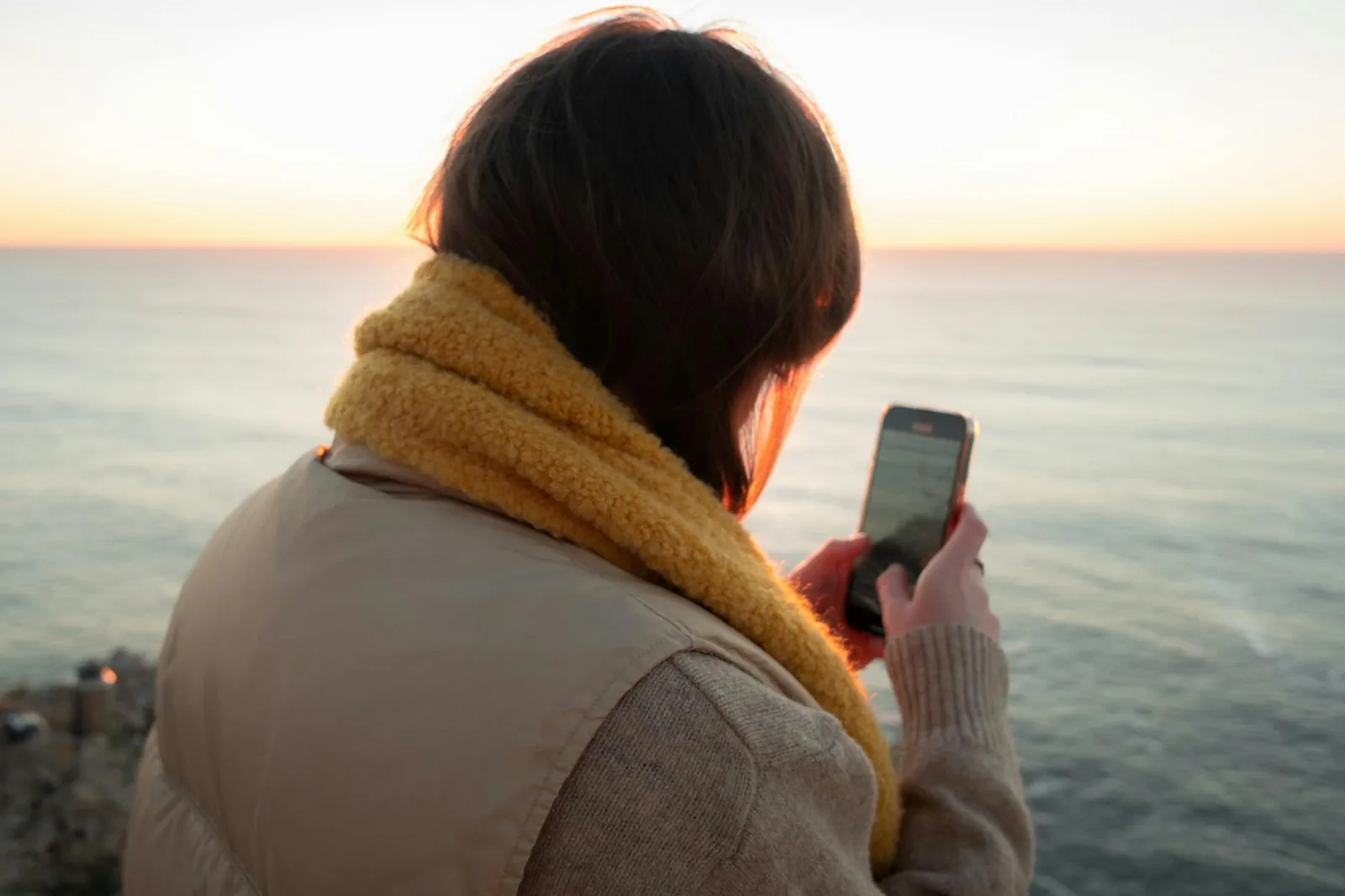 Woman using a smartphone at sunset by the sea, feeling serene and connected.