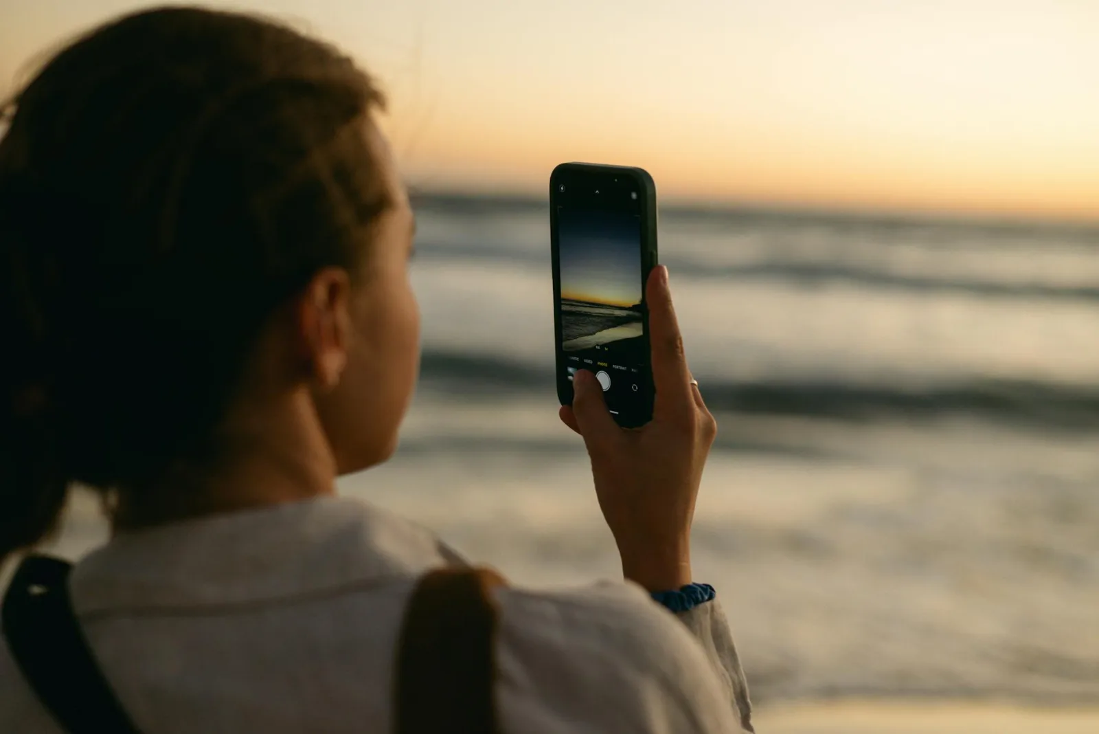 A woman photographing the ocean sunset using her smartphone, showcasing a serene beach atmosphere.
