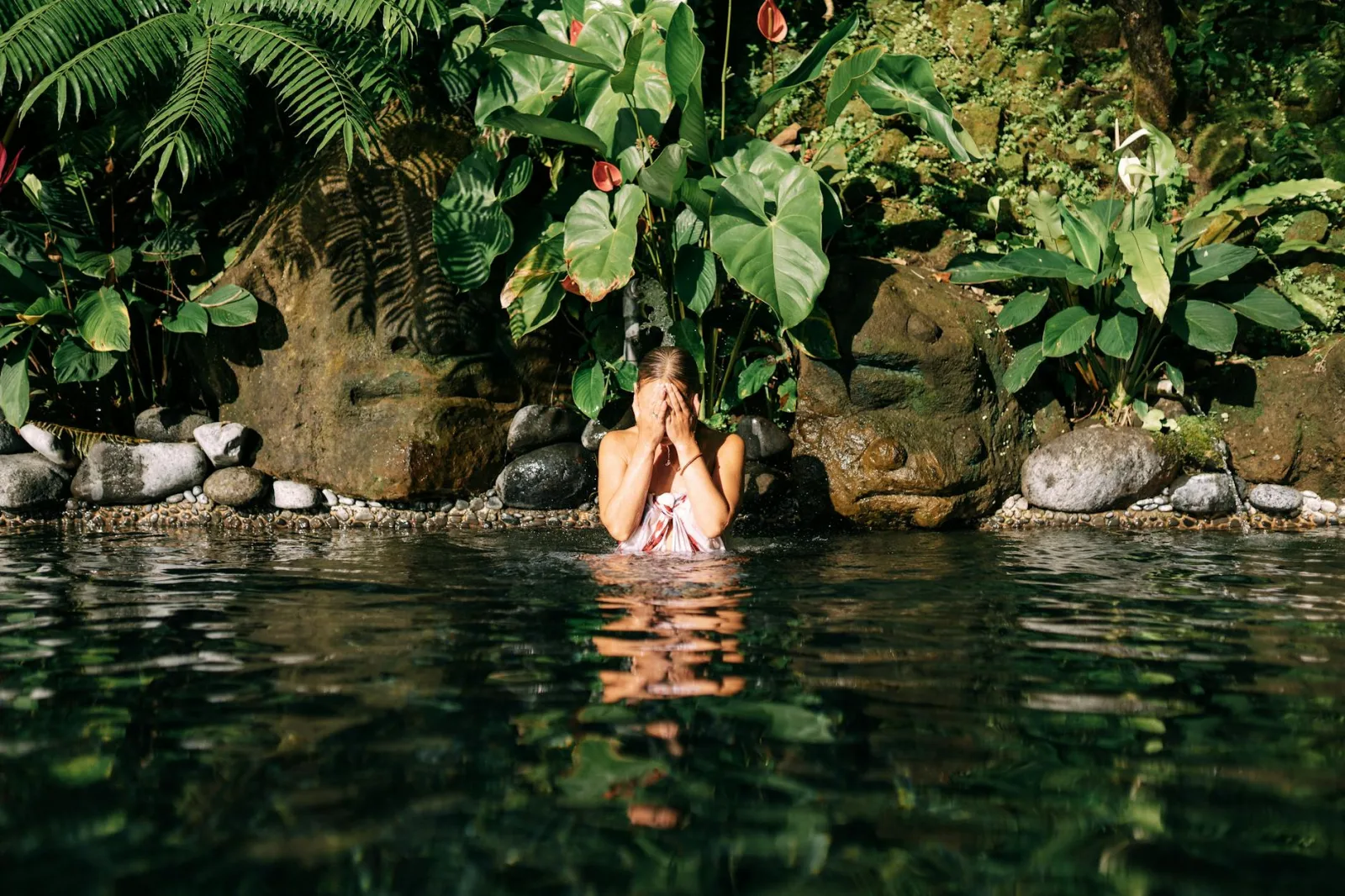 A woman relaxes in a tropical pool surrounded by lush plants and stones.