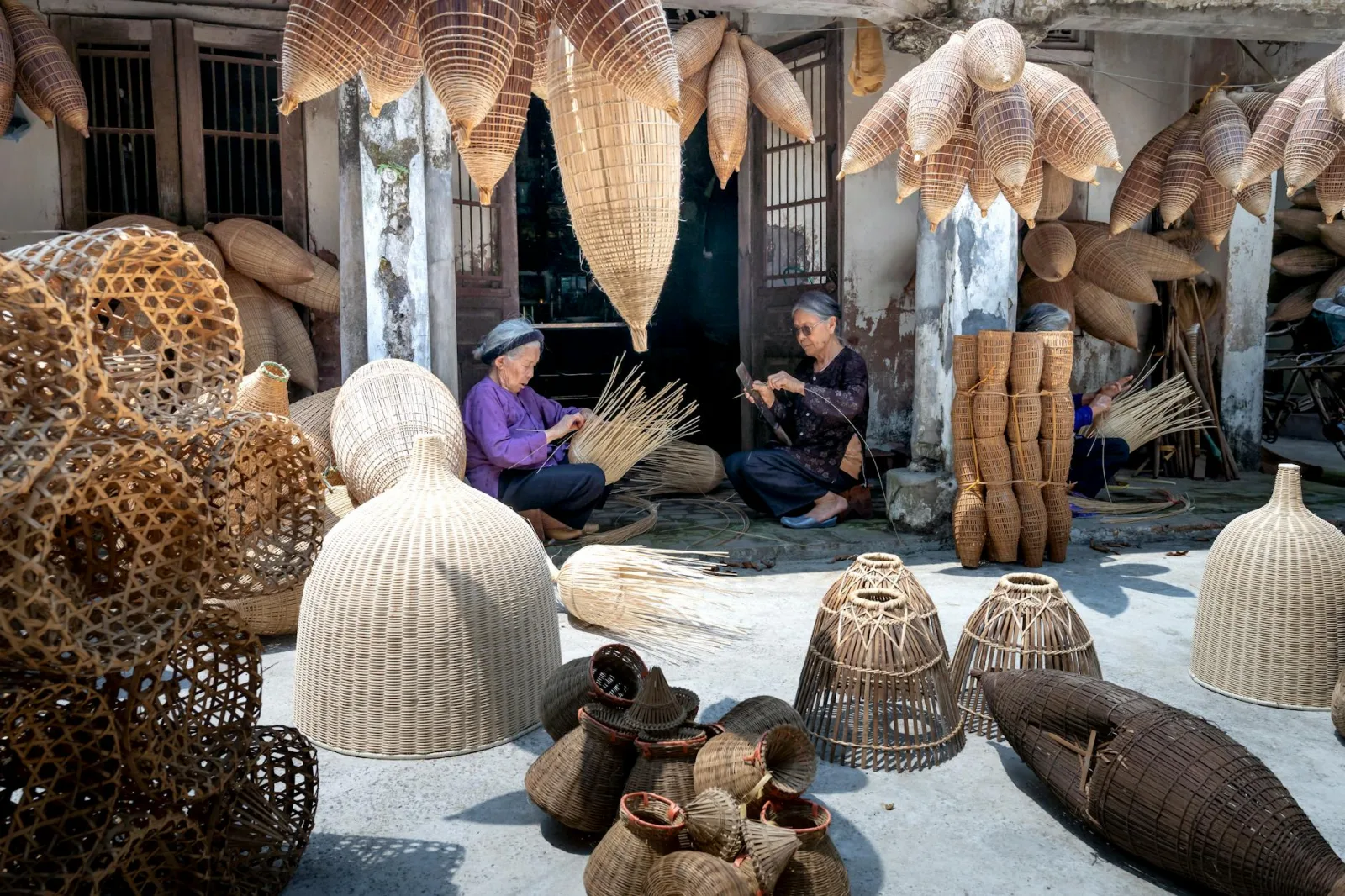 Aged ethnic women making bamboo traditional Vietnamese fish traps placed on ground and hanging in yard in sunlight